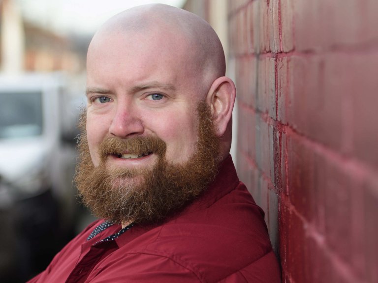 A profile picture of Chris Vaughan, owner of Chris Vaughan Photography. Chris is wearing a red shirt, and is photographed near a red brick wall.