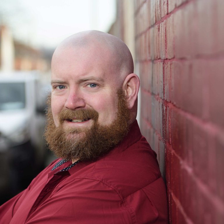 A profile picture of Chris Vaughan, owner of Chris Vaughan Photography. Chris is wearing a red shirt, and is photographed near a red brick wall.