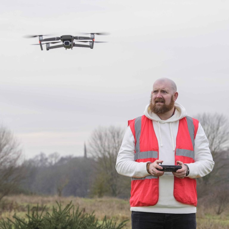 A profile picture of Chris Vaughan, owner of Chris Vaughan Photography, flying a drone. Chris is wearing a white hoodie and a high-vis jacket that has drone pilot written on the back.