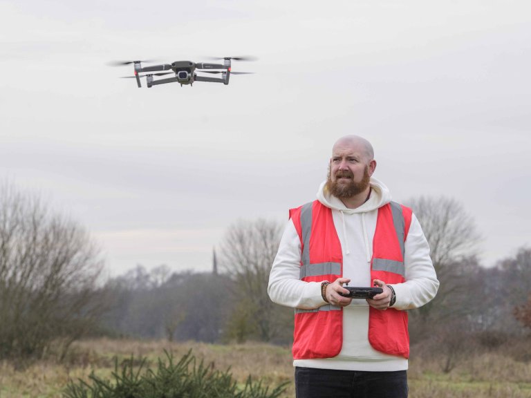 A profile picture of Chris Vaughan, owner of Chris Vaughan Photography, flying a drone. Chris is wearing a white hoodie and a high-vis jacket that has drone pilot written on the back.