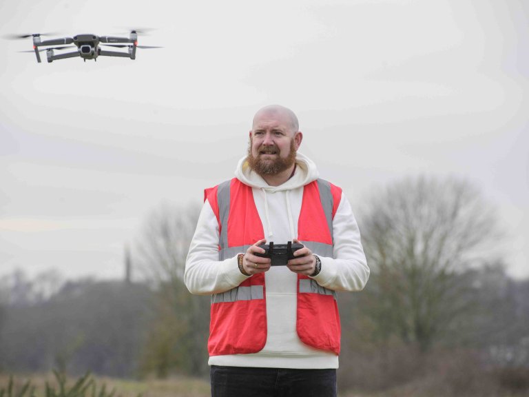 A profile picture of Chris Vaughan, owner of Chris Vaughan Photography, flying a drone. Chris is wearing a white hoodie and a high-vis jacket that has drone pilot written on the back.