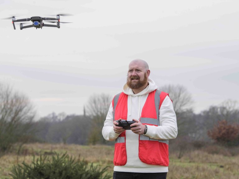 A profile picture of Chris Vaughan, owner of Chris Vaughan Photography, flying a drone. Chris is wearing a white hoodie and a high-vis jacket that has drone pilot written on the back.
