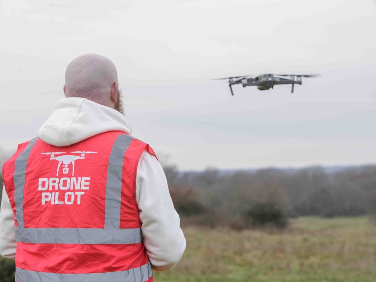 A profile picture of Chris Vaughan, owner of Chris Vaughan Photography, flying a drone. Chris is wearing a white hoodie and a high-vis jacket that has drone pilot written on the back.